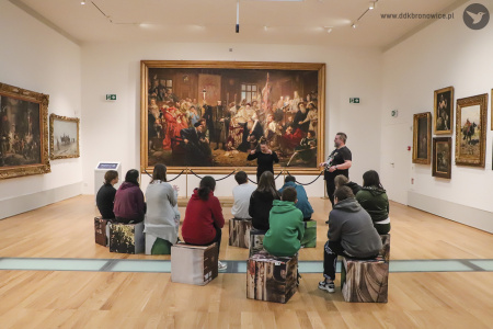 Kolorowe zdjęcie. Grupa młodzieży siedzi tyłem na kwadratowych klockach w sali muzealnej. Przed nimi na ścianie wisi obraz Unii Lubelskiej Jana Matejki. Po prawej stronie od obrazu stoi przewodnik, obok niego miga tłumaczka.