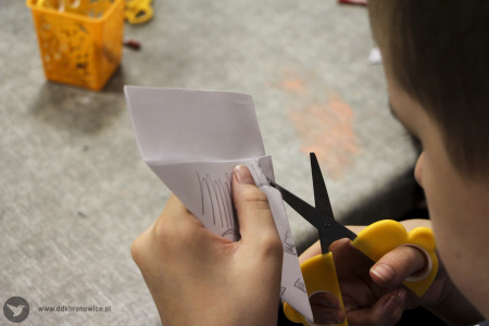 Kolorowe zdjęcie. Chłopięce dłonie trzymają nożyczki tnąc kawałek papieru złożony w rożek.