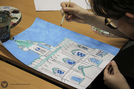 Kolorowe zdjęcie. Na stole leży kartka papieru z naszkicowaną Wieżą Trynitarską. Dziewczęca dłoń trzyma pędzel i maluje farbami szkic.