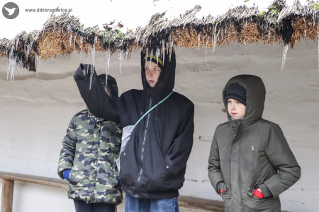 Kolorowe zdjęcie. Trzech chłopców ubranych w kurtki stoi po słomianym daszkiem. Jeden z nich stojący w środku dłonią sięga po zwisający jeden z sopli lodu.