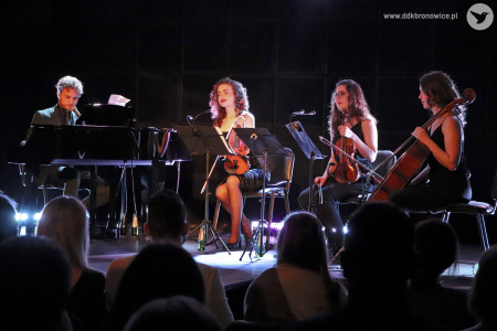 Zdjęcie. Na pierwszym planie głowy publiczności. Na scenie Janek Traczyk gra na fortepianie i śpiewa, obok siedzą skrzypaczki i wiolonczelistka.