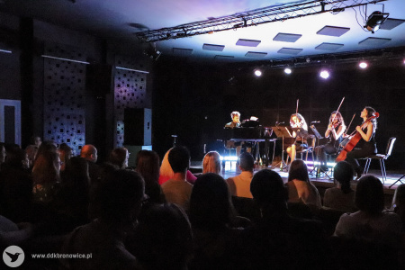 Zdjęcie. Zasłuchana publiczność patrzy na scenę. Na scenie Janek Traczyk i akompaniamentujące mu kobiety wykonują utwór.