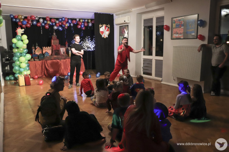 Zdjęcie. Dzieci siedzą na podłodze tyłem w zaciemnieniu. Przodem do nich zwróceni są dwaj aktorzy. Ubrany na czerwono mężczyzna wyciąga dłonie w kierunku dzieci. Drugi mężczyzna stoi z tyłu z założonymi rękami.