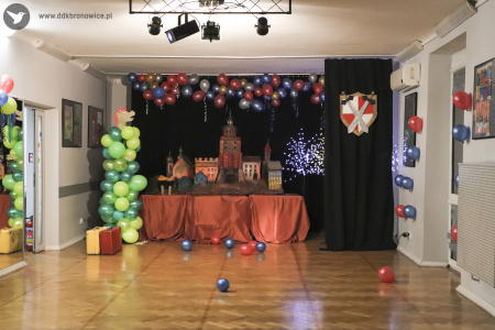 Zdjęcie. Na sali w centrum na podwyższeniu stoi makieta zamku. Z lewej strony stoi smok wykonany z balonów. W tle wiszą balony. Po prawej stronie na zasłonie powieszona jest tarcza z mieczami.