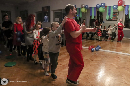 Zdjęcie. Dzieci tańczą w wężyku. Prowadzi ich aktor w czerwonym ubraniu. W tle widać lustro i ich odbicia.