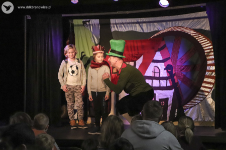 Zdjęcie. Na scenie stoją aktor oraz dwójka dzieci z publiczności. Aktor mówi do dzieci. Na drugim planie tło przedstawiające młyn.