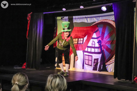 Zdjęcie. Na scenie stoi aktor. Ma na sobie wysoki, zielony kapelusz. Stoi w dynamicznej pozie i zwraca się do publiczności. na drugim planie tło przedstawiające młyn.