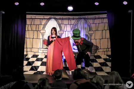 Zdjęcie. Aktorka w pomarańczowej sukience trzyma duże czerwone spodnie. Aktor w zielonym kapeluszu ogląda je. Na drugim planie tło przedstawiające ścianę z oknami, widzianą od wewnątrz.