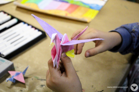 Kolorowe zdjęcie. Dziewczynka składa origami przy stoliku.