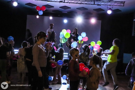 Kolorowe zdjęcie. Na zaciemnionej sali grupa dzieci, w tle kobieta z kolorowymi balonami.
