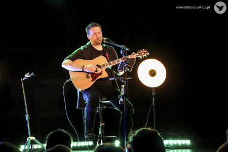 Kolorowe zdjęcie. Na ciemnej scenie Michał Łangowski siedzi na wysokim krześle, gra na gitarze i śpiewa. Przed nim pulpit na nuty. W tle ciemnej sceny lampa dająca jedno punktowe światło.