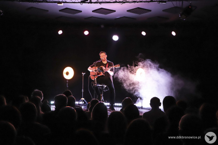 Kolorowe zdjęcie. Na ciemnej scenie Michał Łangowski siedzi na wysokim krześle, gra na gitarze i śpiewa. Przed nim widownia. Za nim unosi się dym sceniczny.