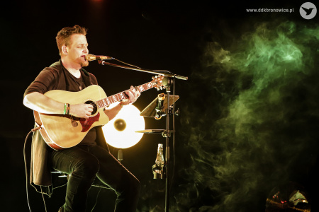 Kolorowe zdjęcie. Na ciemnej scenie Michał Łangowski siedzi na wysokim krześle, gra na gitarze i śpiewa. W tle lampa i dym sceniczny.