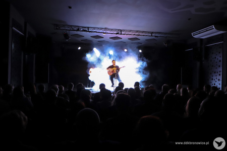 Kolorowe zdjęcie. Kadr zza pleców publiczności. W tle Michał Łangowski na oświetlonej scenie gra na gitarze.