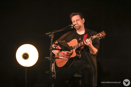 Kolorowe zdjęcie. Na ciemnej scenie Michał Łangowski siedzi na wysokim krześle, gra na gitarze i śpiewa.