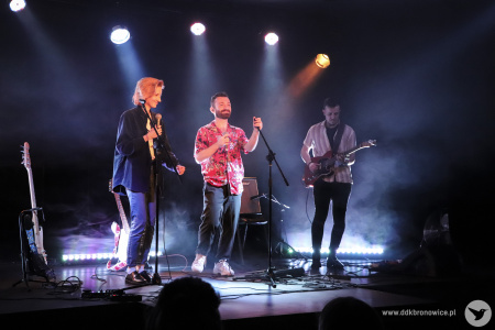 Kolorowe zdjęcie. Na scenie Aga, Adrian i Piotrek. Aga i Adrian trzymają mikrofony w dłoniach. Piotrek gra na gitarze.