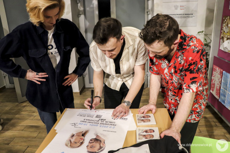 Kolorowe zdjęcie. Aga, Adrian i Piotrek podpisują plakat koncertu.