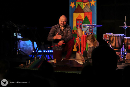 Kolorowe zdjęcie. Na scenie Marek Fedor i dziewczynka. Muzyk klęczy, przed nim są cymbały. Dziewczynka gra pałeczkami na cymbałach.