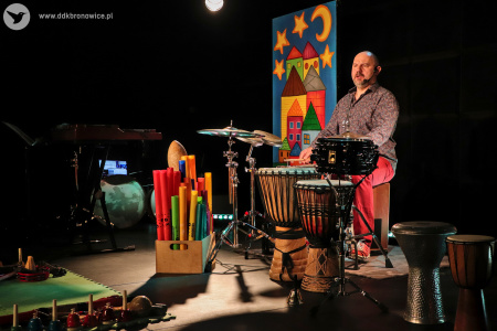Kolorowe zdjęcie. Na scenie Marek Fedor wśród wielu instrumentów muzycznych, takich jak: djembe, bum bum rurki, perkusja. Muzyk gra na perkusji.