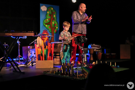 Kolorowe zdjęcie. Na scenie Marek Fedor z chłopcem. Chłopiec gra pałeczkami na perkusji.