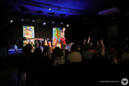 Kolorowe zdjęcie. Kadr na scenę zza publiczności. Marek Fedor na scenie uśmiecha się w stronę widzów. Kika osób podnosi ręce w geście zgłaszania się.