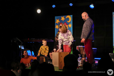 Kolorowe zdjęcie. Na scenie Marek Fedor z dwójką dzieci. Dziewczynka i chłopiec grają na kahonach.