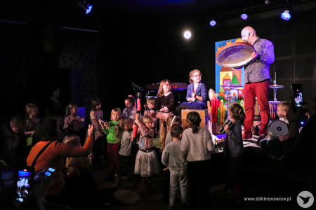 Kolorowe zdjęcie. Na scenie grupa dzieci z Markiem Fedorem. Muzyk gra na bębnie obręczowym. Dzieci wpatrują się w Marka.