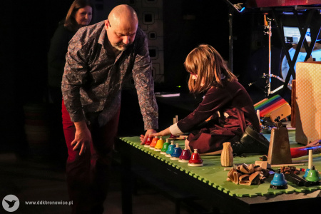 Kolorowe zdjęcie. Na scenie dziewczynka gra na kolorowych dzwonkach. Przed sceną Marek Fedor przygląda się jak dziewczynka gra.