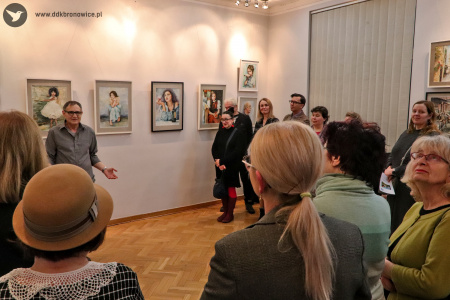 Kolorowe zdjęcie. Grupa osób patrzy na Wiesława Stefańskiego, który stoi na tle swoich obrazów.