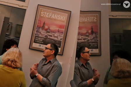 Kolorowe zdjęcie. Kadr na Wiesława Stefańskiego z profilu. Jego odbicie widać w telewizorze. Artysta stoi obok plakatu promującego wystawę.