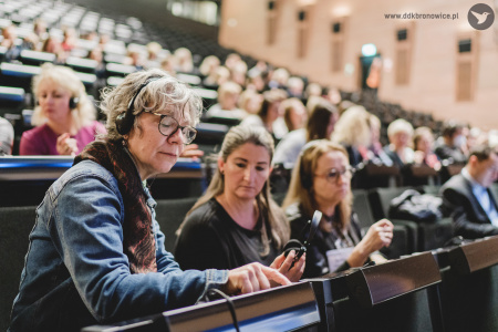 Kolorowe zdjęcie. Audytorium w Europejskim Centrum Solidarności w Gdańsku. Uczestnicy konferencji siedzą na widowni audytorium. Część z nich ma słuchawki na uszy. Na pierwszym planie Kobieta w słuchawkach i okularach z zamkniętymi oczami wskazuje palcem na pulpit przed sobą.