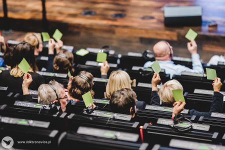 Kolorowe zdjęcie. Zdjęcie uchwycone z góry od tyłu. Uczestnicy konferencji siedzą na widowni w słuchawkach i unoszą zielone kartki w dłoni do góry.