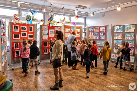 Kolorowe zdjęcie. Grupa dzieci i dwie kobiety oglądają prace plastyczne w Galerii Małej. Nad nimi wiszą witrażowe anioły.