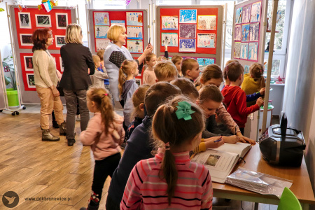 Kolorowe zdjęcie. Grupa dzieci ogląda prace plastyczne w Galerii Małej. Niektóre dzieci podpisują się w księdze pamiątkowej. Kobieta fotografuje dzieci telefonem.