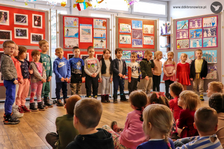 Kolorowe zdjęcie. Grupa dzieci stoi w rzędzie na tle wystawy. Przed nimi na podłodze siedzą inne dzieci zwrócone w ich kierunku.