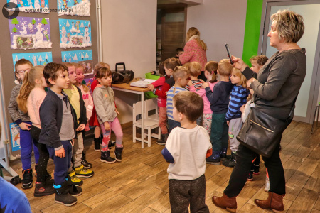 Kolorowe zdjęcie. Grupa dzieci pozuje na tle prac plastycznych w Galerii Małej. Kobieta fotografuje dzieci telefonem.