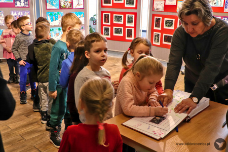 Kolorowe zdjęcie. Dzieci stoją w kolejce do księgi pamiątkowej. Jedna dziewczynka wpisuje się do księgi z pomocą nauczycielki.