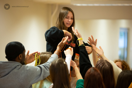Kolorowe zdjęcie. Uczestnicy warsztatów zgromadzeni wokół stojącej wyżej dziewczyny. Kierują ku niej swoje ręce z zawiązanymi kolorowymi sznurkami.