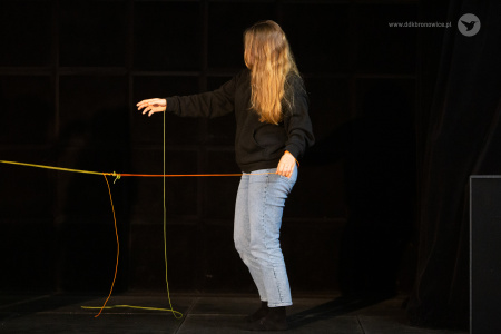 Kolorowe zdjęcie. Dziewczyna stoi bokiem na scenie, z tyłu czarne tło.  Prawą dłoń z przywiązanym żółtym sznurkiem kieruje do przodu. Lewą dłonią ciągnie pomarańczowy sznurek, związany z żółtym.