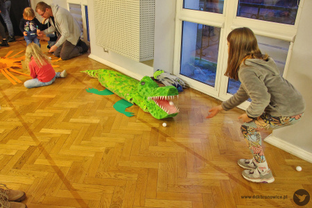Kolorowe zdjęcie. Dziewczynka rzuca piłeczką do paszczy tekturowego krokodyla.