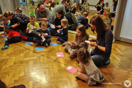 Kolorowe zdjęcie. Grupa dzieci i dorosłych siedzi na podłodze na tle lustra. Dzieci wycinają zwierzątka z kolorowej pianki.