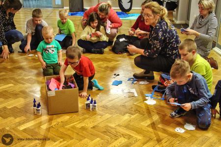 Kolorowe zdjęcie. Grupa dzieci siedzi na podłodze i wycina zwierzątka z kolorowej pianki. Jeden chłopiec sięga do kartonu z materiałami plastycznymi.