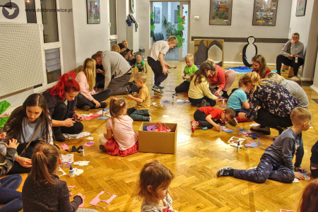 Kolorowe zdjęcie. Grupa dzieci i dorosłych siedzi na podłodze. Dzieci wycinają zwierzątka z kolorowej pianki. Niektórzy dorośli pomagaja dzieciom w pracy plastycznej.