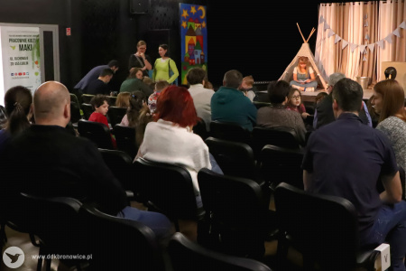 Kolorowe zdjęcie. Kadr na salę kameralną w Pracowniach Kultury Maki. Widok na plecy ludzi siedzących na krzesłach. W tle scena z bezowymi dekoracjami.