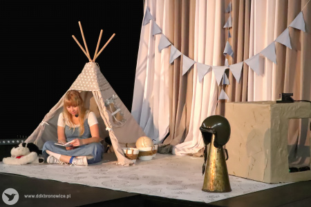 Kolorowe zdjęcie. Aktorka siedzi po turecku na scenie w małym namiocie tipi i czyta książkę. W tle beżowo-szare dekoracje: kurtyna, girlanda.