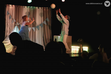 Kolorowe zdjęcie. Aktorzy na scenie odbijają do siebie piłkę. Mają ręce wyciągnięte do góry, jakby grali w piłkę siatkową.