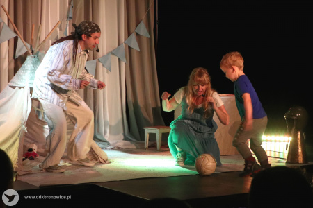 Kolorowe zdjęcie. Na scenie mały chłopiec i aktorzy. Chłopiec bierze rozbieg, aby kopnąć piłkę w stronę mężczyzny. Kobieta dopinguje chłopca.