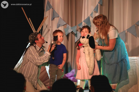 Kolorowe zdjęcie. Na scenie aktorzy, chłopiec i dziewczynka. Aktorzy trzymają blisko dzieci pluszowe misie.