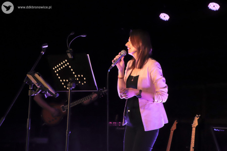 Kolorowe zdjęcie. Na scenie skupiona kobieta śpiewa do mikrofonu. Kadr z boku. Za nią, na scenie stoją dwie gitary.