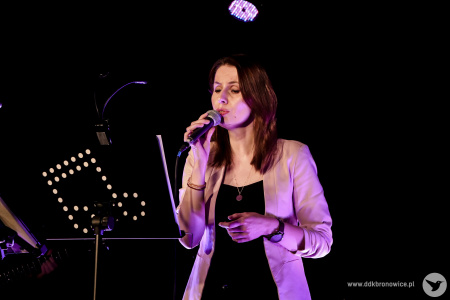 Kolorowe zdjęcie. Na scenie kobieta śpiewa do mikrofonu. Zerka na pulpit do nut.